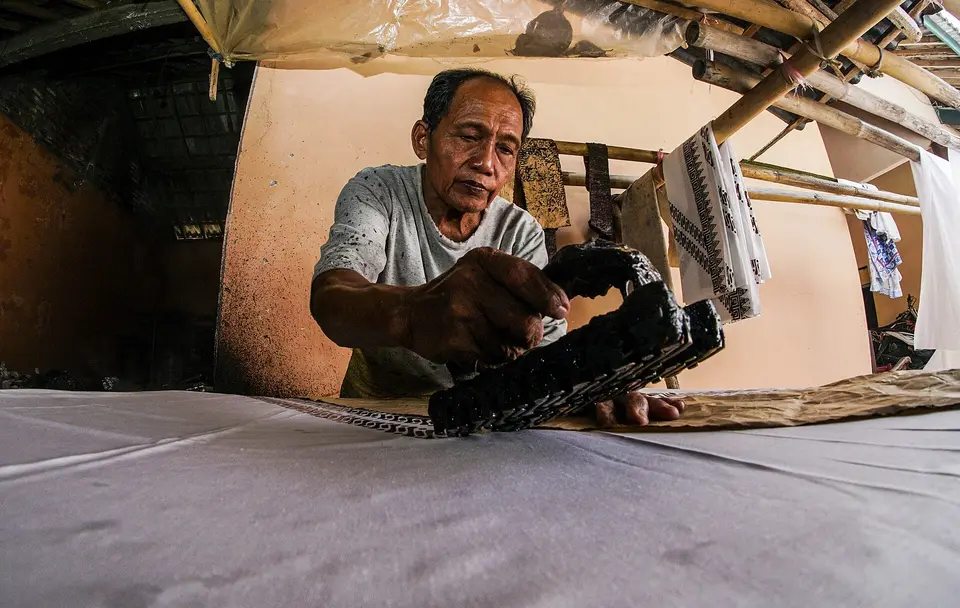 Perajin pria mencapkan cap tembaga bermalam ke atas kain yang terbentang di meja untuk membuat batik cap